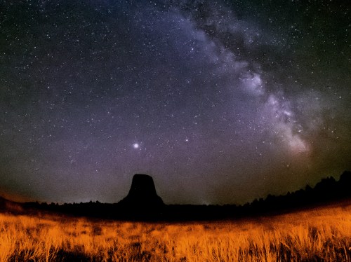 A starry night sky with the Milky Way over a dark silhouette of a rock formation in a grassy landscape.