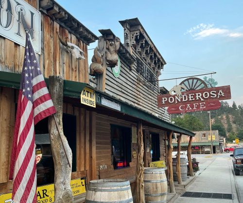 A rustic wooden café with a sign reading "Ponderosa Café," featuring barrels and an American flag outside.
