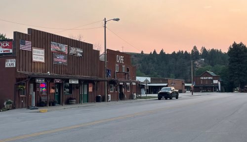 A quiet street in a small town at sunset, featuring rustic buildings and a few parked vehicles.