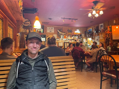 A man sits on a bench in a rustic restaurant, with diners and decor visible in the background.
