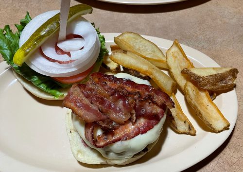 A bacon cheeseburger with melted cheese, lettuce, tomato, and onion, served with golden fries on a plate.