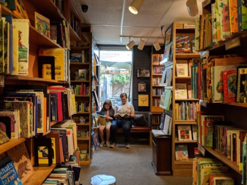 A cozy bookstore aisle filled with colorful books, featuring two people sitting and reading in a sunlit corner.