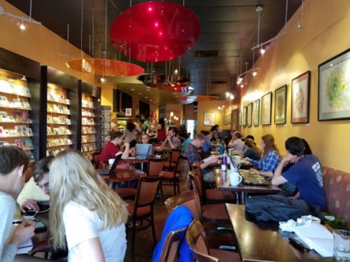 A busy café with patrons at tables, some working on laptops, surrounded by colorful artwork and bookshelves.