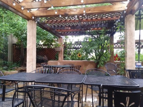 A cozy outdoor patio with black metal tables and chairs, surrounded by greenery and string lights.