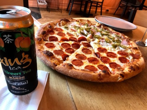 A large pizza topped with pepperoni, green peppers, and onions, next to a can of mango green tea on a wooden table.