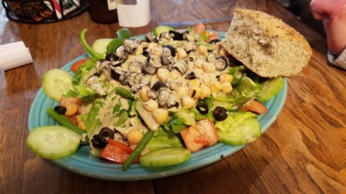 A colorful salad with greens, tomatoes, cucumbers, black olives, chickpeas, and a piece of bread on the side.