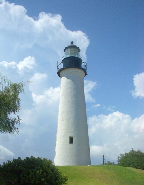 A tall white lighthouse stands on a grassy hill under a blue sky with fluffy clouds.