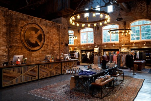 A stylish retail store with exposed brick walls, wooden displays, and warm lighting, showcasing clothing and accessories.