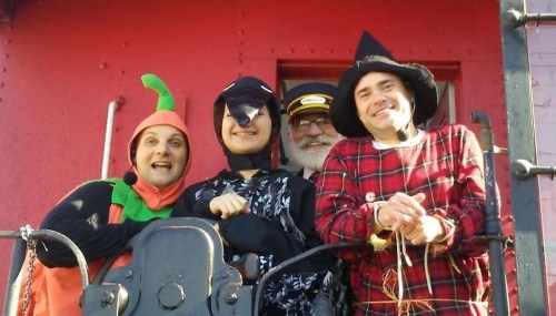 Four people in costumes, including a pumpkin, witch, and a bird, smiling together on a train platform.