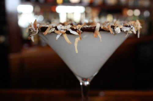 A close-up of a cocktail in a martini glass, garnished with shredded coconut and chocolate on the rim.