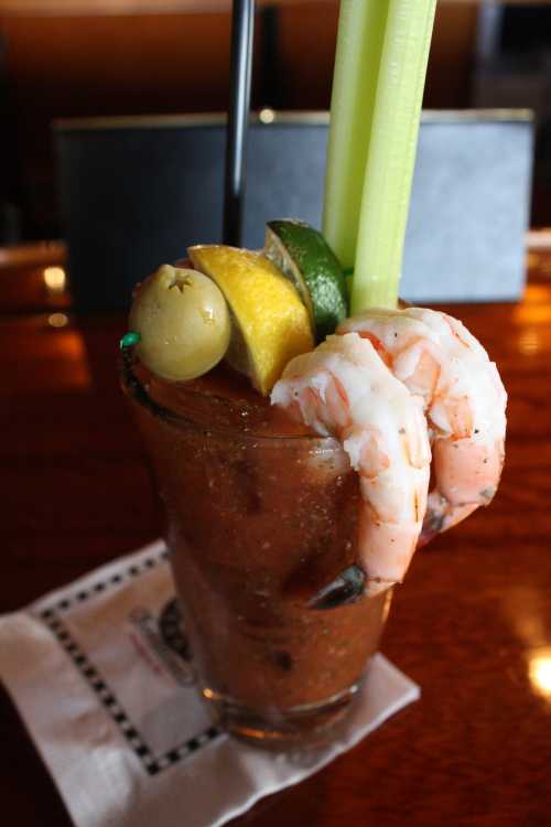 A tall glass of Bloody Mary garnished with shrimp, celery, olives, and lime on a wooden bar counter.