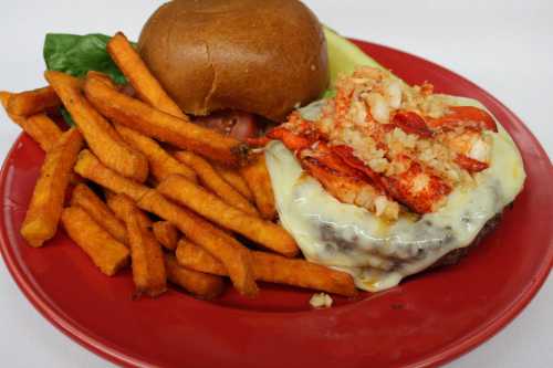 A plate featuring a cheeseburger topped with lobster, served with sweet potato fries and a bun.