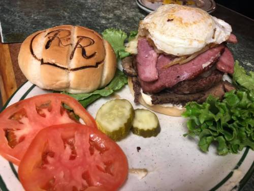 A stacked burger with lettuce, tomatoes, pickles, and a fried egg, served on a plate with a toasted bun.