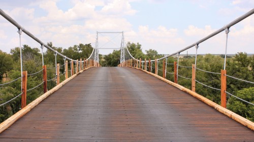 A wooden suspension bridge stretches over a landscape, surrounded by trees under a partly cloudy sky.