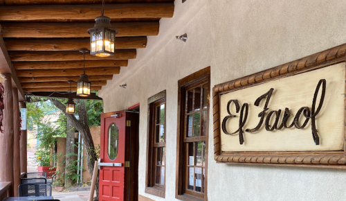 A rustic building exterior with a wooden sign reading "El Farol" and hanging lanterns under a wooden beam ceiling.