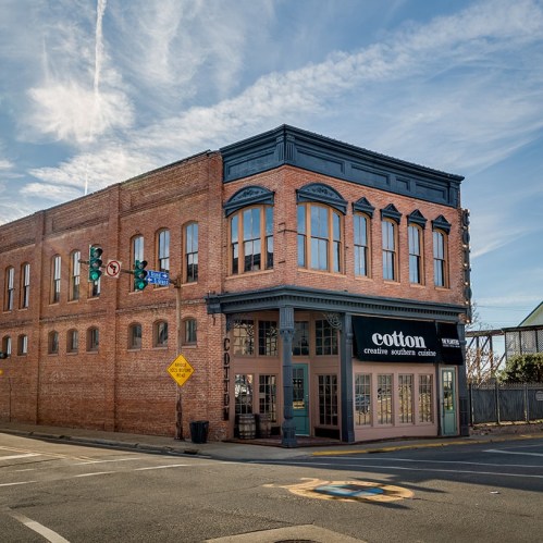 Historic brick building with large windows, featuring a "cotton" restaurant sign at the corner of a street.