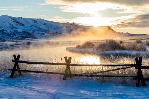 A serene winter landscape with a misty river, snow-covered mountains, and a sunrise illuminating the scene.