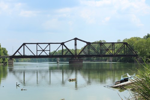 A rusted railway bridge spans a calm river, reflecting the surrounding greenery and blue sky. A small boat is moored nearby.