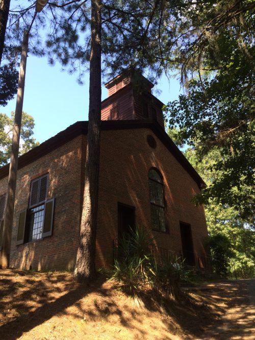 A historic brick building with a bell tower, surrounded by tall trees and greenery, set on a sloped path.