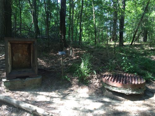An old metal box and a rusty grate sit in a wooded area, surrounded by trees and underbrush.