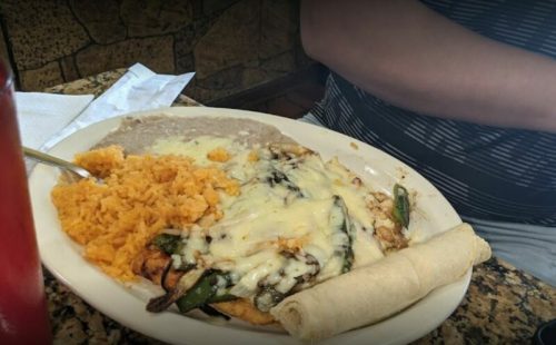 A plate of Mexican food featuring cheese-covered grilled vegetables, rice, refried beans, and a tortilla.