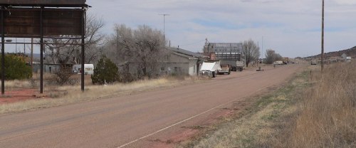 Deserted road with sparse vegetation, old buildings, and a few vehicles under a cloudy sky.