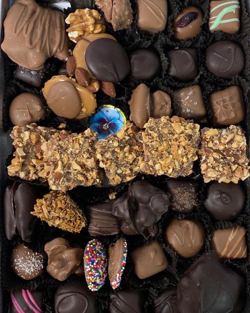 A variety of chocolates and confections arranged in a box, featuring nuts, sprinkles, and a blue flower.