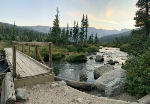 A wooden bridge over a stream, surrounded by trees and mountains under a colorful sky at dusk.
