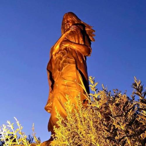 A tall statue of a figure with long hair, standing amidst greenery, illuminated by soft evening light.