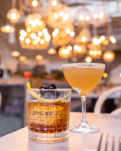 Two cocktails on a table, one with cherries and lemon, the other with a salted rim, surrounded by warm, glowing lights.