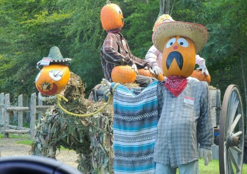 A whimsical display of scarecrows with pumpkin heads, dressed in colorful outfits, on a wooden cart in a green setting.