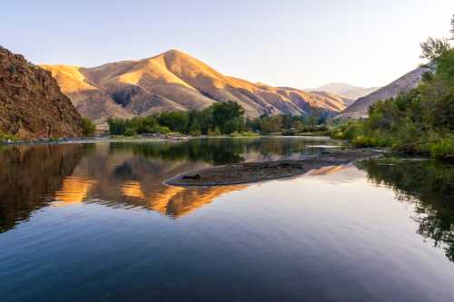 Serene river scene with mountains reflecting in the water at sunset, surrounded by lush greenery.