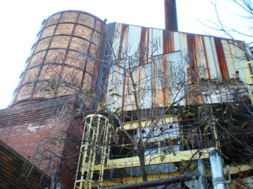 Rusty industrial structure with a tall cylindrical tank, surrounded by bare branches and a blue sky.