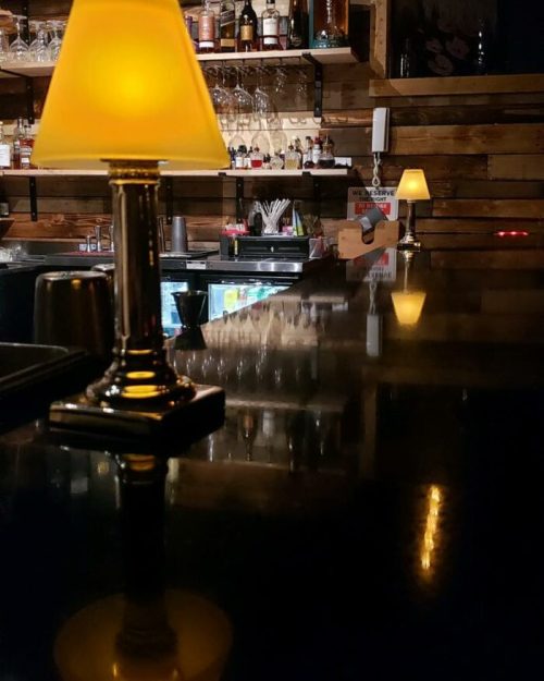 A dimly lit bar scene featuring a small lamp on the counter, with shelves of bottles in the background.