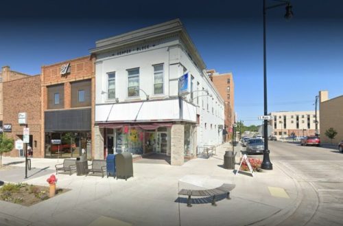 A street view of a corner building with a storefront, benches, and nearby buildings in an urban setting.