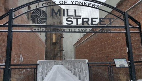 Snow-covered entrance to Mill Street in Yonkers, featuring a metal archway and a walkway leading into a narrow alley.