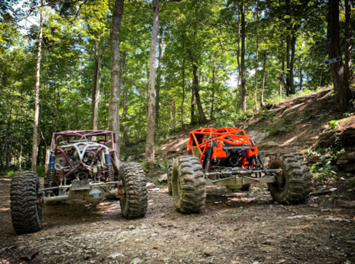 Two off-road vehicles parked in a wooded area, surrounded by trees and rocky terrain.