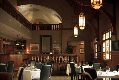 Elegant restaurant interior with tables set for dining, wooden accents, and stained glass windows. Warm, inviting atmosphere.