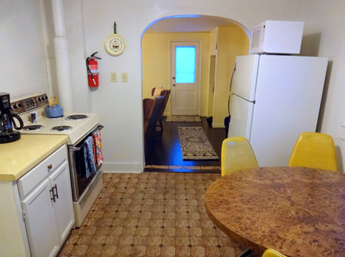 A cozy kitchen with a stove, refrigerator, and a round table with yellow chairs, leading to a door with a blue window.