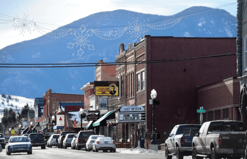 A snowy street scene with shops and cars, framed by mountains and festive decorations overhead.