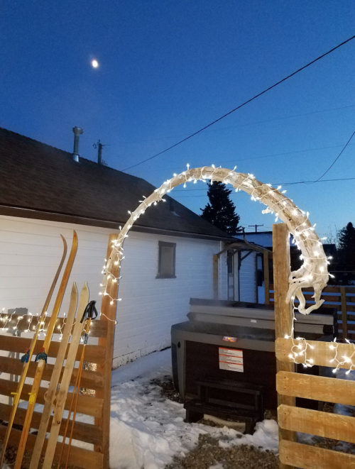 A snowy scene with a lit archway, skis leaning against a wooden fence, and a clear evening sky with a crescent moon.