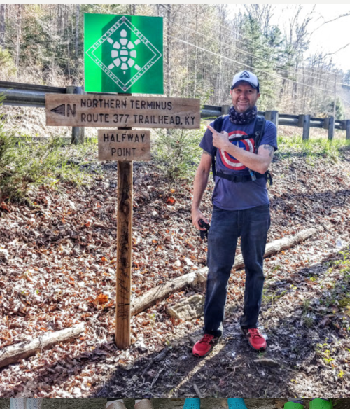 A man stands next to a trail sign indicating the northern terminus and halfway point on Route 377 in Kentucky.