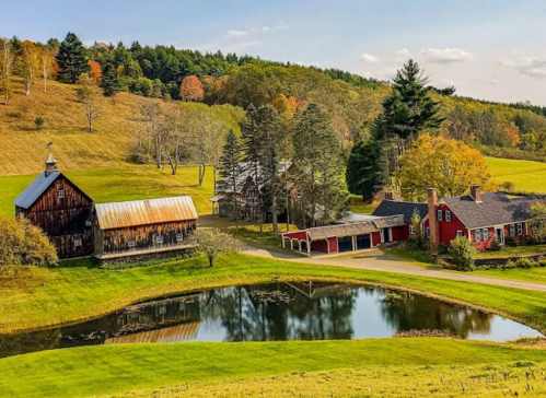 A scenic view of a farm with a pond, red barn, and autumn foliage on rolling hills under a clear sky.