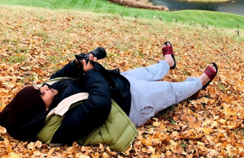 A person lies on a bed of autumn leaves, holding a camera, wearing a jacket, sweatpants, and sandals.