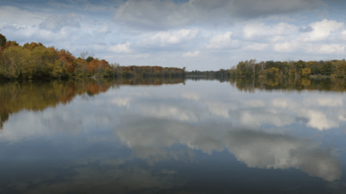 A calm lake reflecting trees and clouds, surrounded by autumn foliage under a partly cloudy sky.