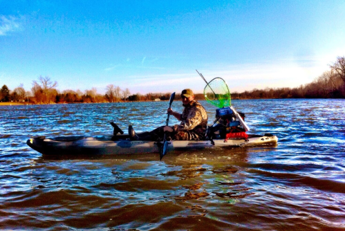 A person paddles a kayak on a river, equipped with fishing gear and a net, under a clear blue sky.