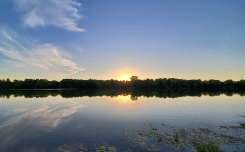 A serene sunset over a calm lake, with reflections of trees and clouds in the water.