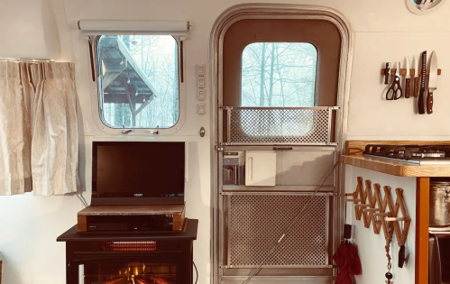 Cozy interior of a camper with a window, a small TV, kitchen tools, and a heater, surrounded by a serene, snowy landscape.