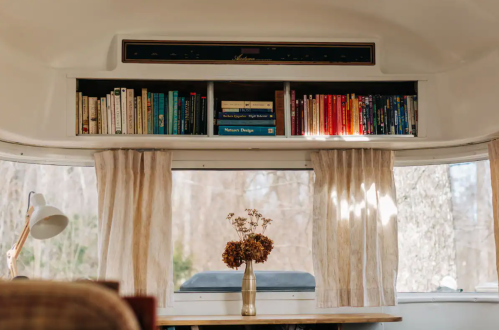 Cozy interior of a camper with bookshelves, a vase of flowers, and soft curtains by a window.