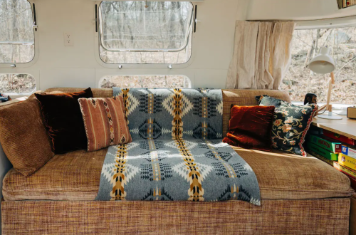 Cozy interior of a camper with a sofa covered in a patterned blanket and various decorative pillows. Natural light filters in.